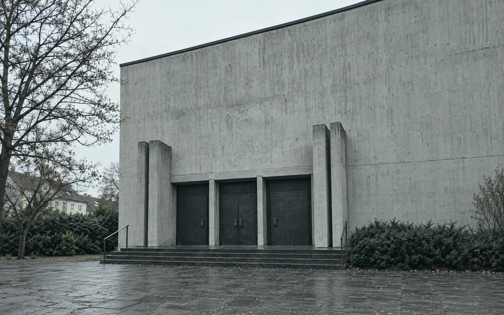 Die schlichte Eingangsseite einer Kirche aus Sichtbeton mit drei dunklen Bronzetüren, die von vier pfeilerartigen Mauerstücken gerahmt werden.