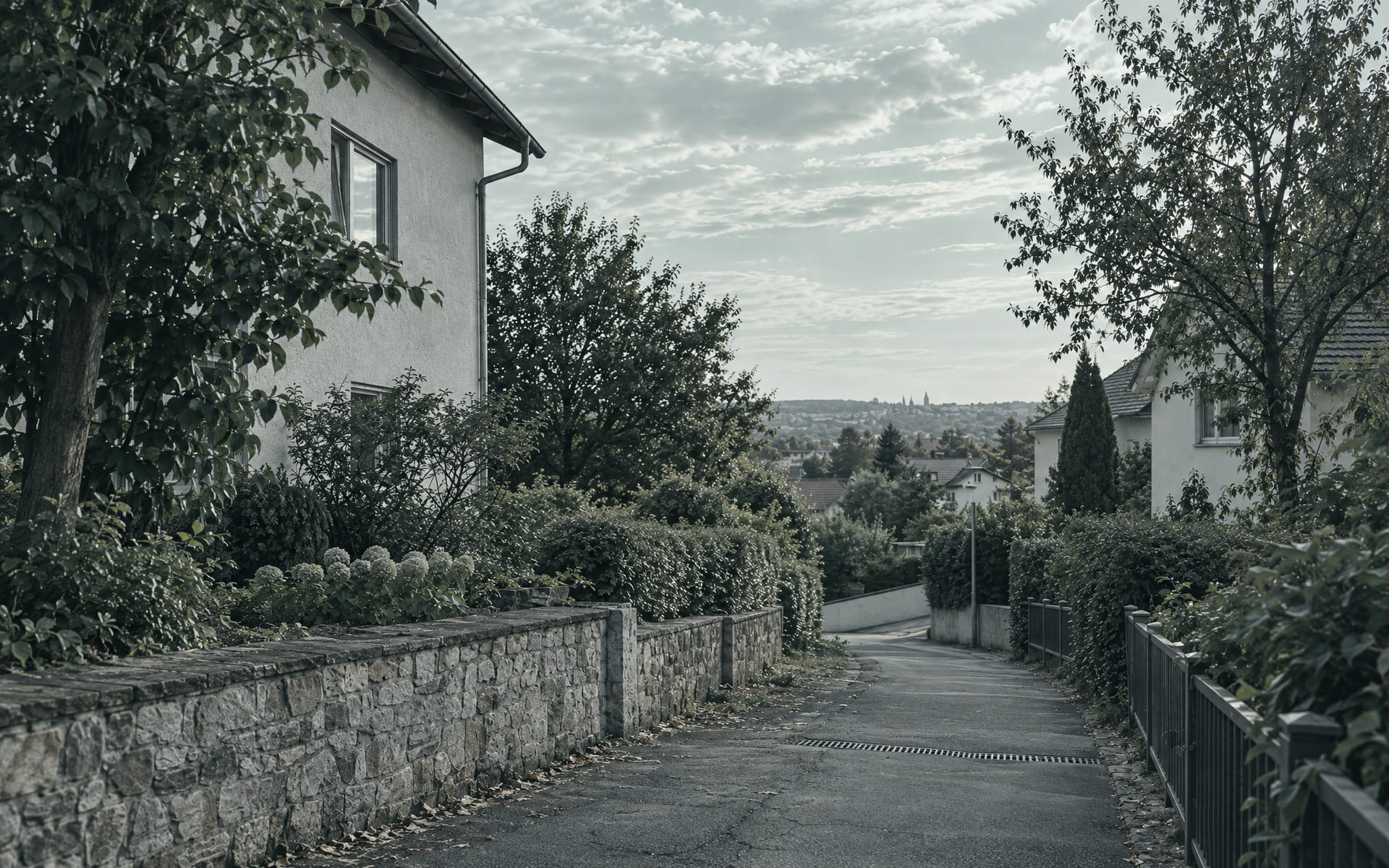 Ein schmaler Wohnweg fällt zwischen einer niedrigen Steinmauer und Hecken leicht ab, im Hintergrund stehen Häuser unter einem bedeckten Himmel.