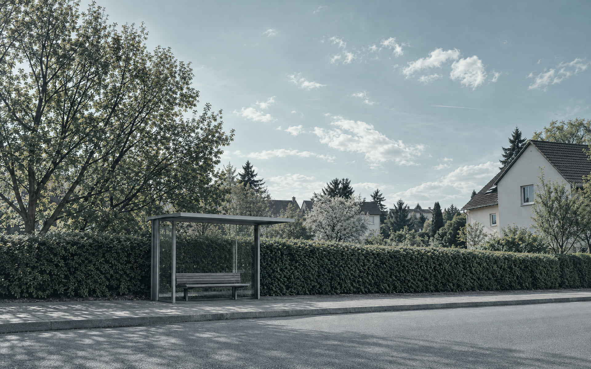 Eine leere Wartehalle an einer Haltestelle im Wohngebiet, dahinter eine Hecke und ein Stück Gehweg im Nachmittagslicht.